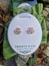 Load image into Gallery viewer, A pair of rose gold shamrock stud earrings with small green gemstone accents, displayed on a white oval card branded 'TRINITY & CO. IRISH JEWELLERY'.